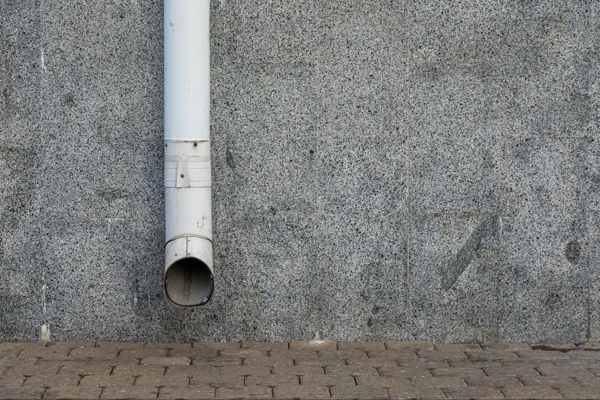 A white downspout attached to a grey exterior wall for rainwater drainage.