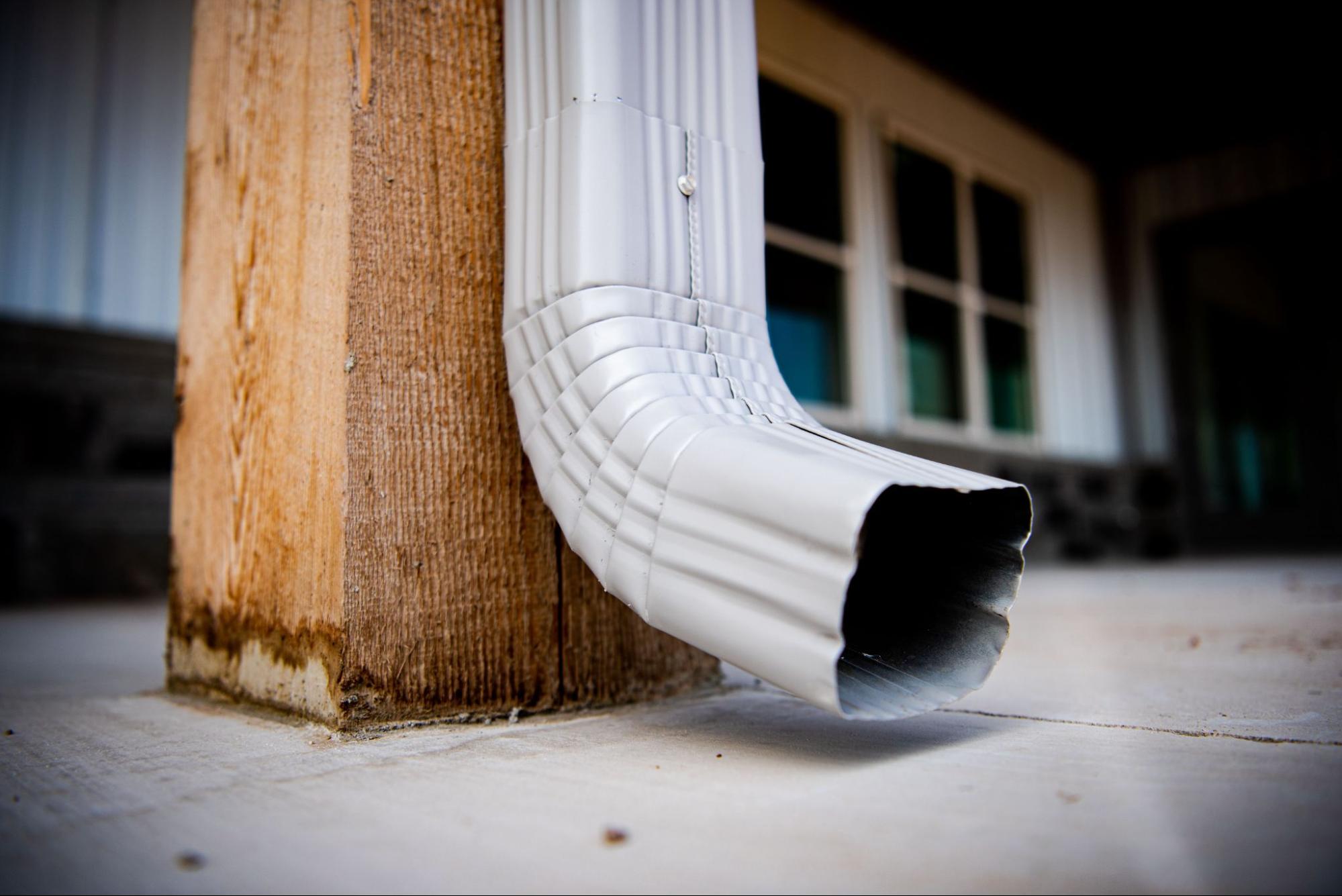 Close-up of a silver square downspout gutter attached to a wooden pole with a house in the background