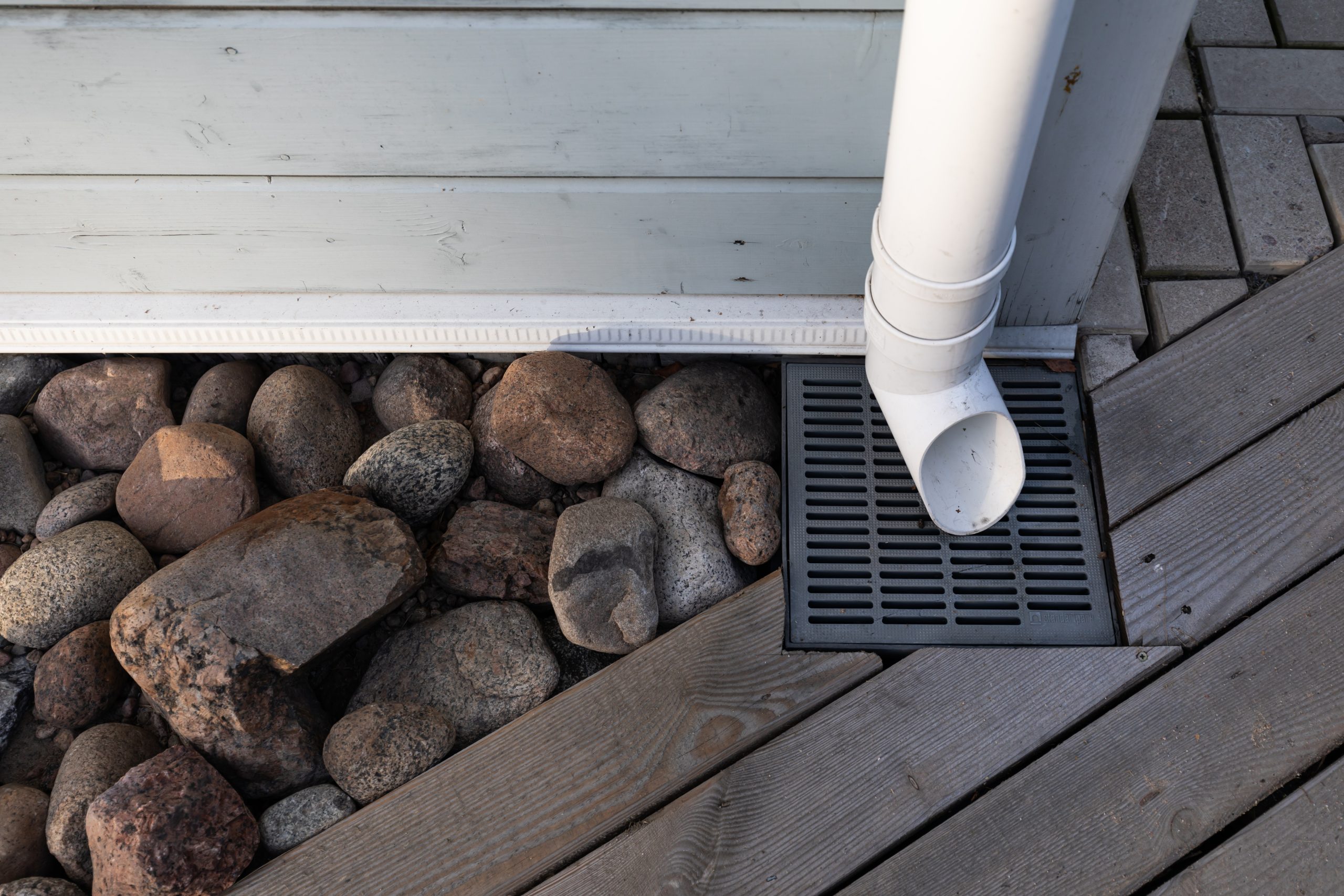 A white rainwater drain pipe and a drainage system with a grate built into the wooden platform and decorated with stones A white rainwater drain pipe and a drainage system with a grate built into the wooden platform and decorated with stones