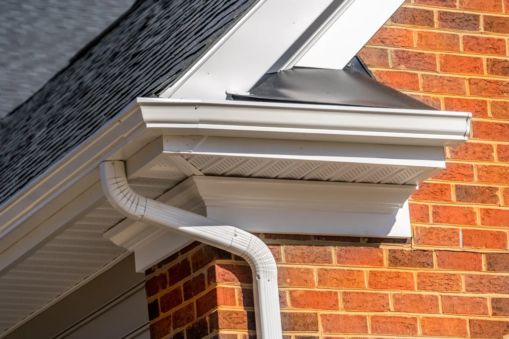 Close-up of white outside gutters and a soffit vent on a house with a brick wall and dark shingle roof