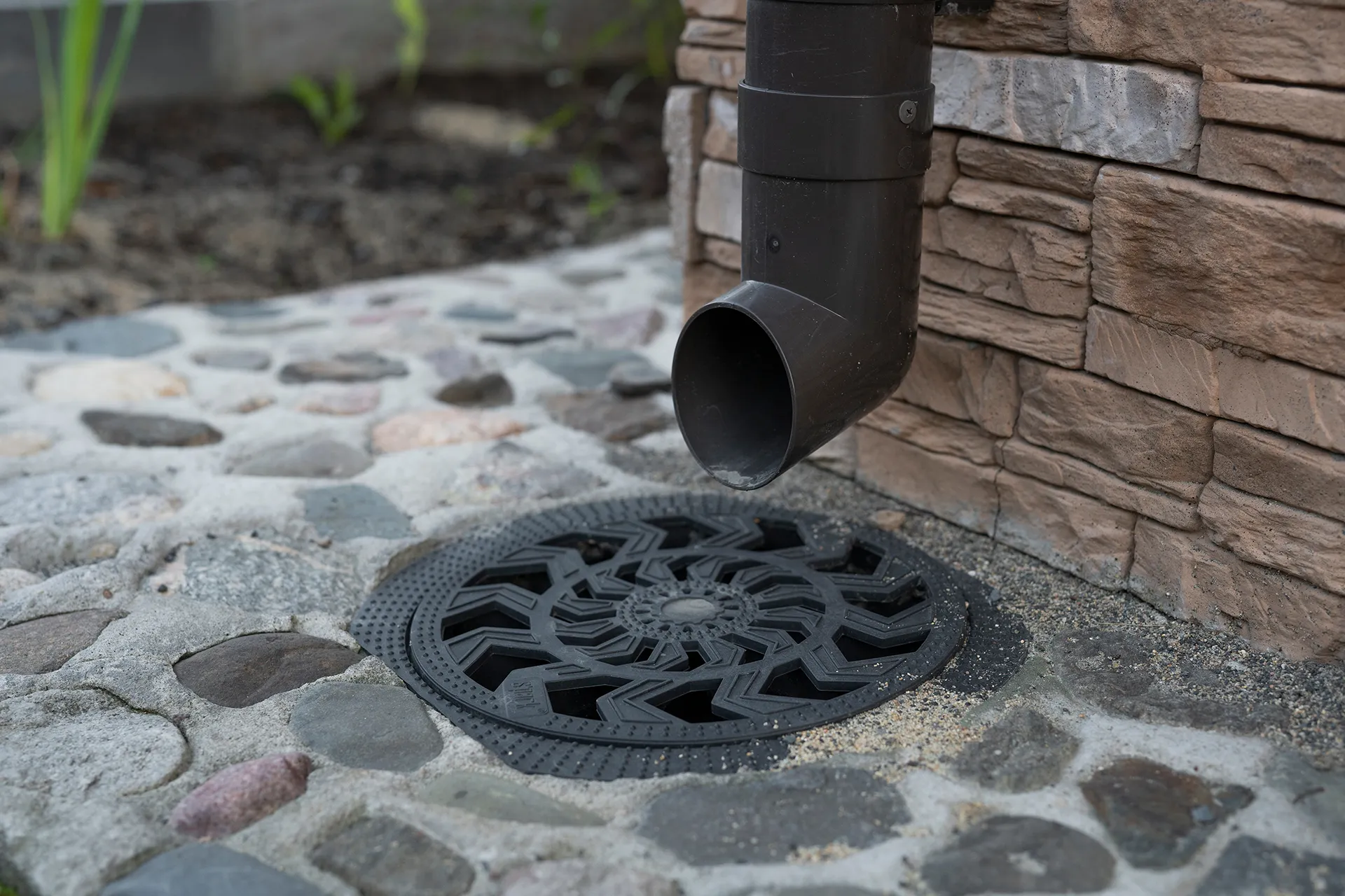 A dark grey downspout emptying directly onto a decorative black circular drain cover set into a cobblestone-like ground, next to a stone-faced wall.