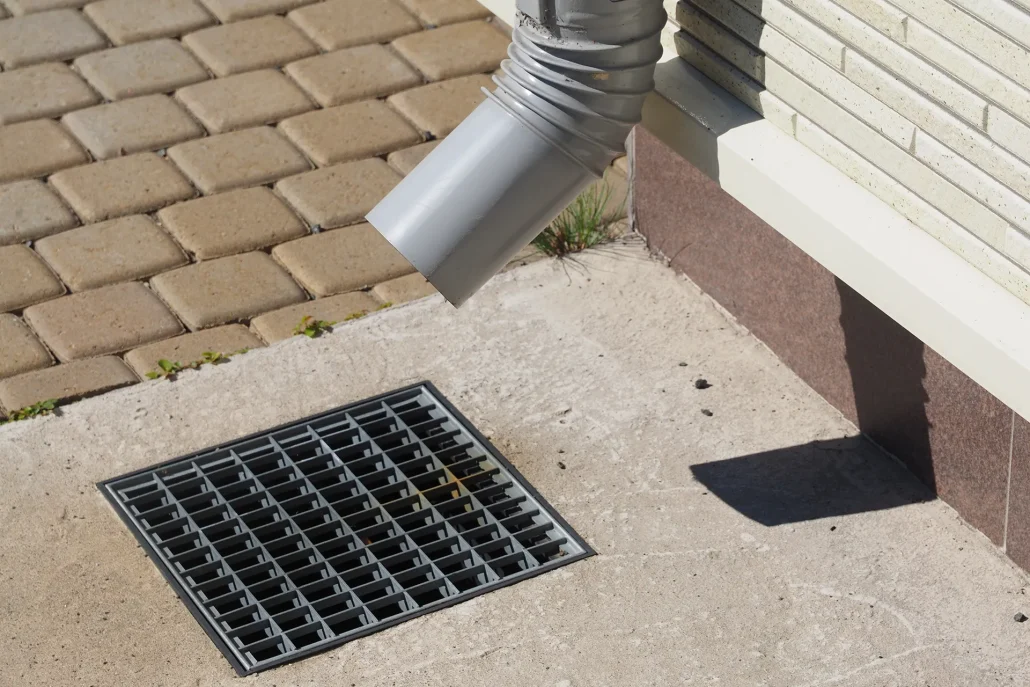 A light grey downspout redirects water away from a light-colored brick wall and brown foundation, emptying near a metal grate drain set into a concrete path, with beige pavers in the background.