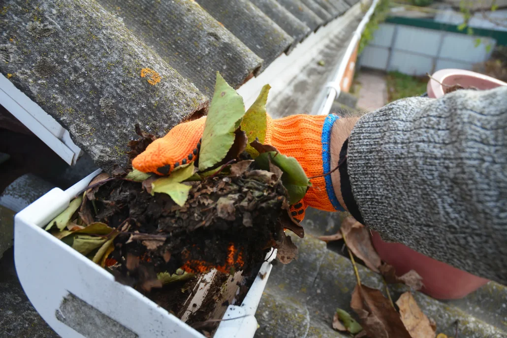 Close-up of a person wearing orange gloves, manually removing a large pile of wet leaves and debris from a dirty house gutter.