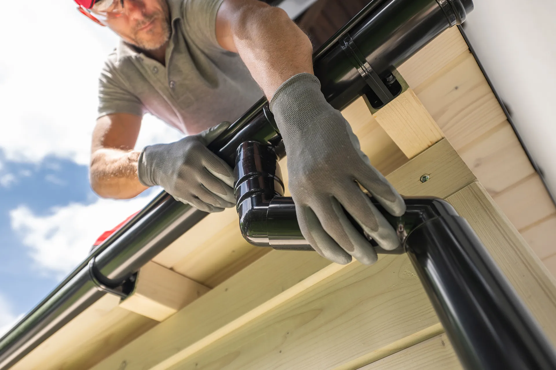Close-up of a worker in gloves installing a black downspout section to a gutter system on wooden eaves, demonstrating part of an eaves protection system.