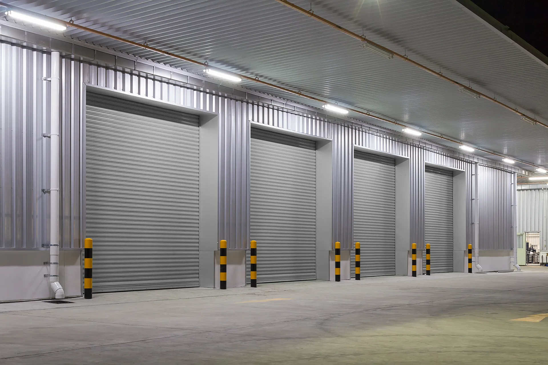 Exterior of an industrial or commercial building featuring multiple grey roll-up garage doors with corrugated metal siding walls, illuminated by overhead lighting at dusk or night.