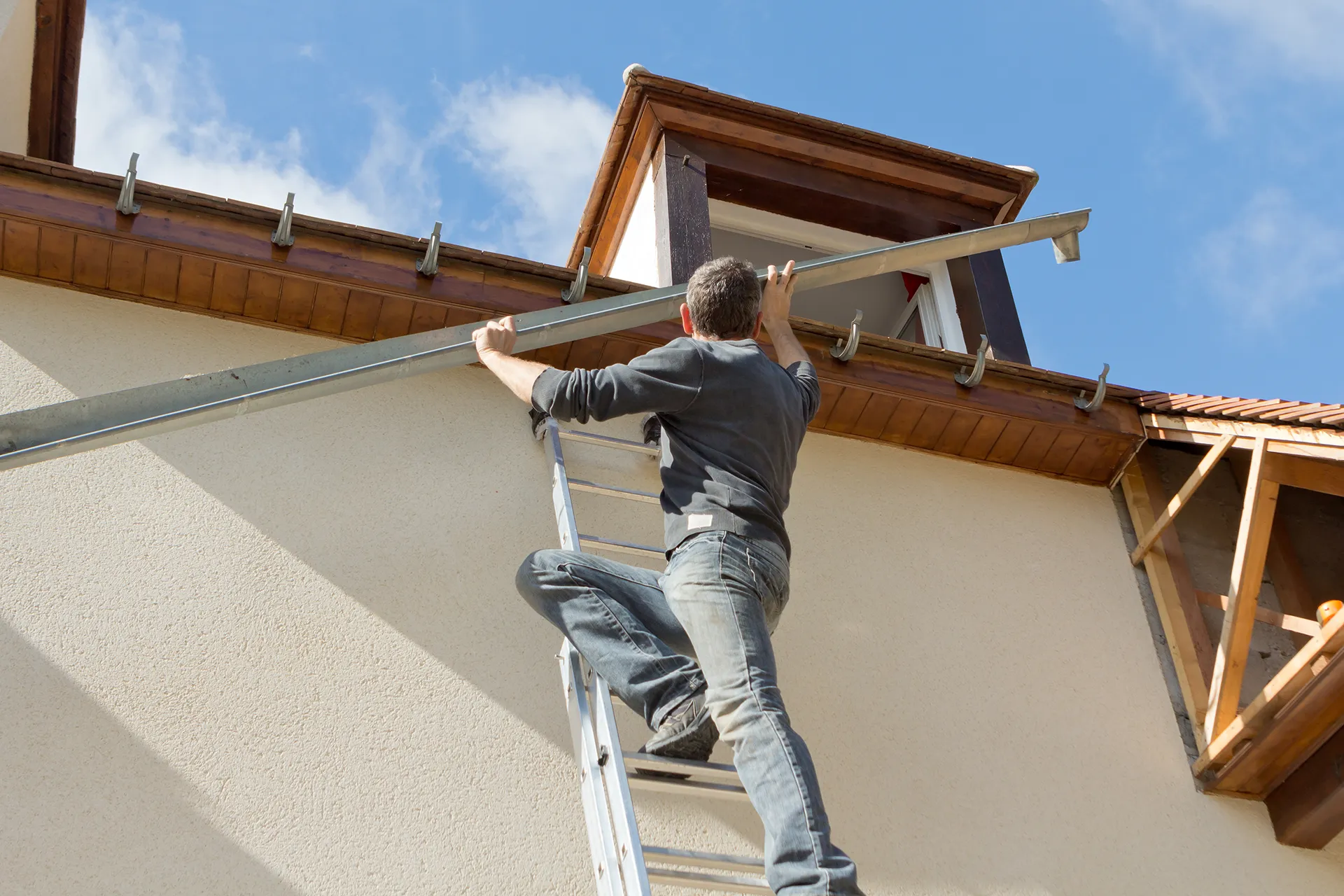 Man on a ladder installing a new section of gutter on a house, attaching it to the fascia board.