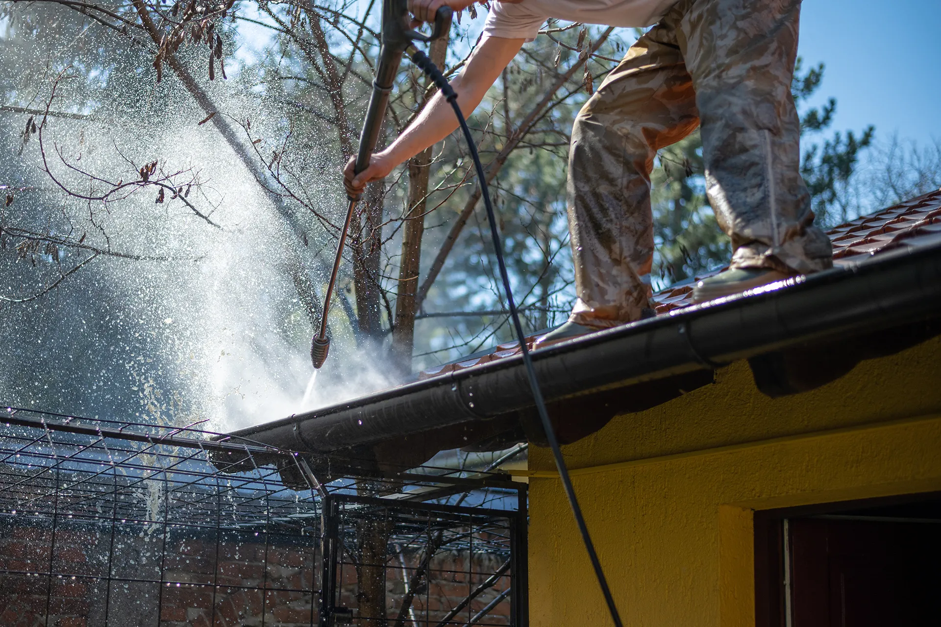 A person wearing work pants and a t-shirt uses a high-pressure washer to clean out dark gutters on the edge of a roof, with water spraying out, demonstrating professional gutter cleaning.