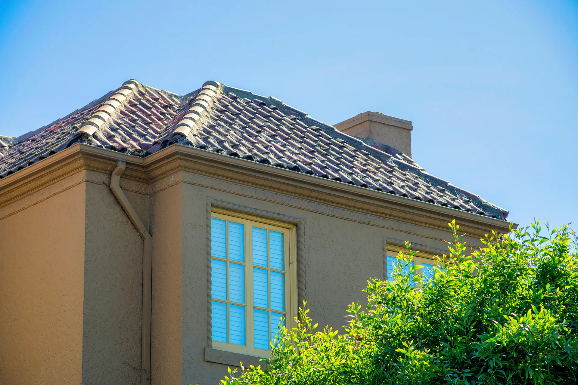 Top portion of a Mediterranean-style stucco house with a red tile roof, beige gutters, and a visible chimney against a clear blue sky.