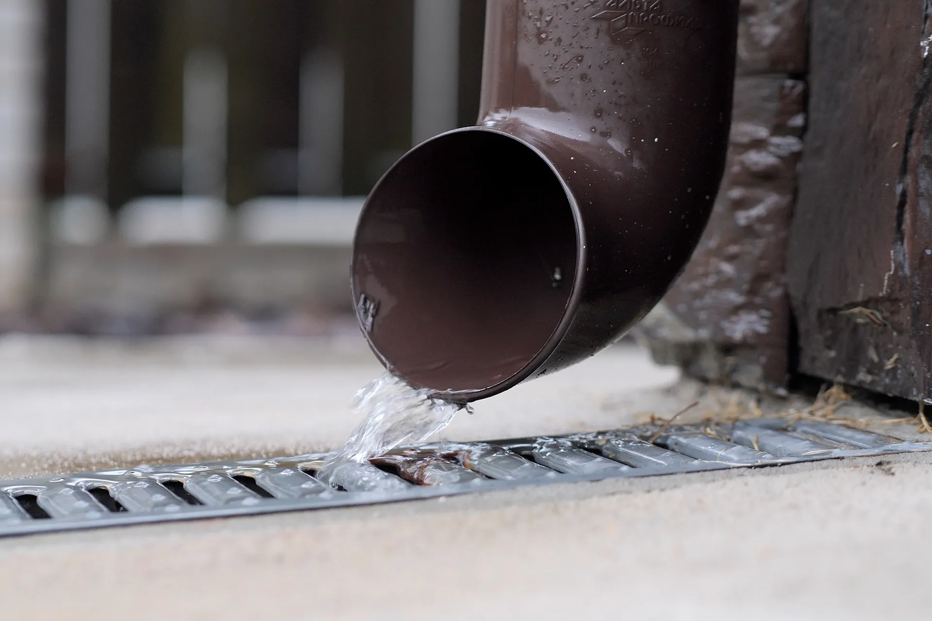Close-up of a brown downspout directing rainwater into a metal grate drain at ground level, with water actively flowing out of the pipe.
