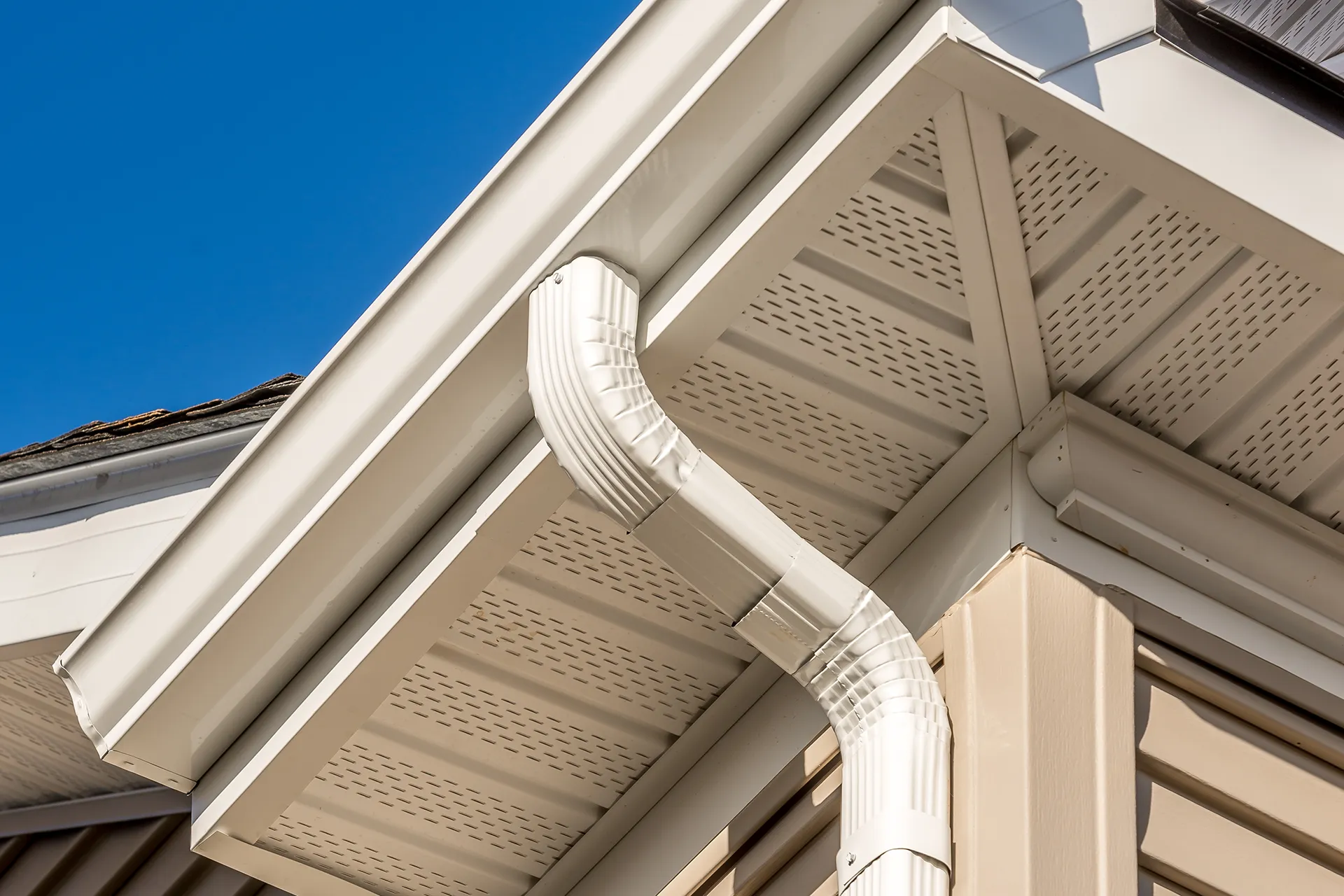 Close-up view of white TaylorMade seamless gutters and downspout on the exterior of a house with beige siding and white soffit, under a clear blue sky.