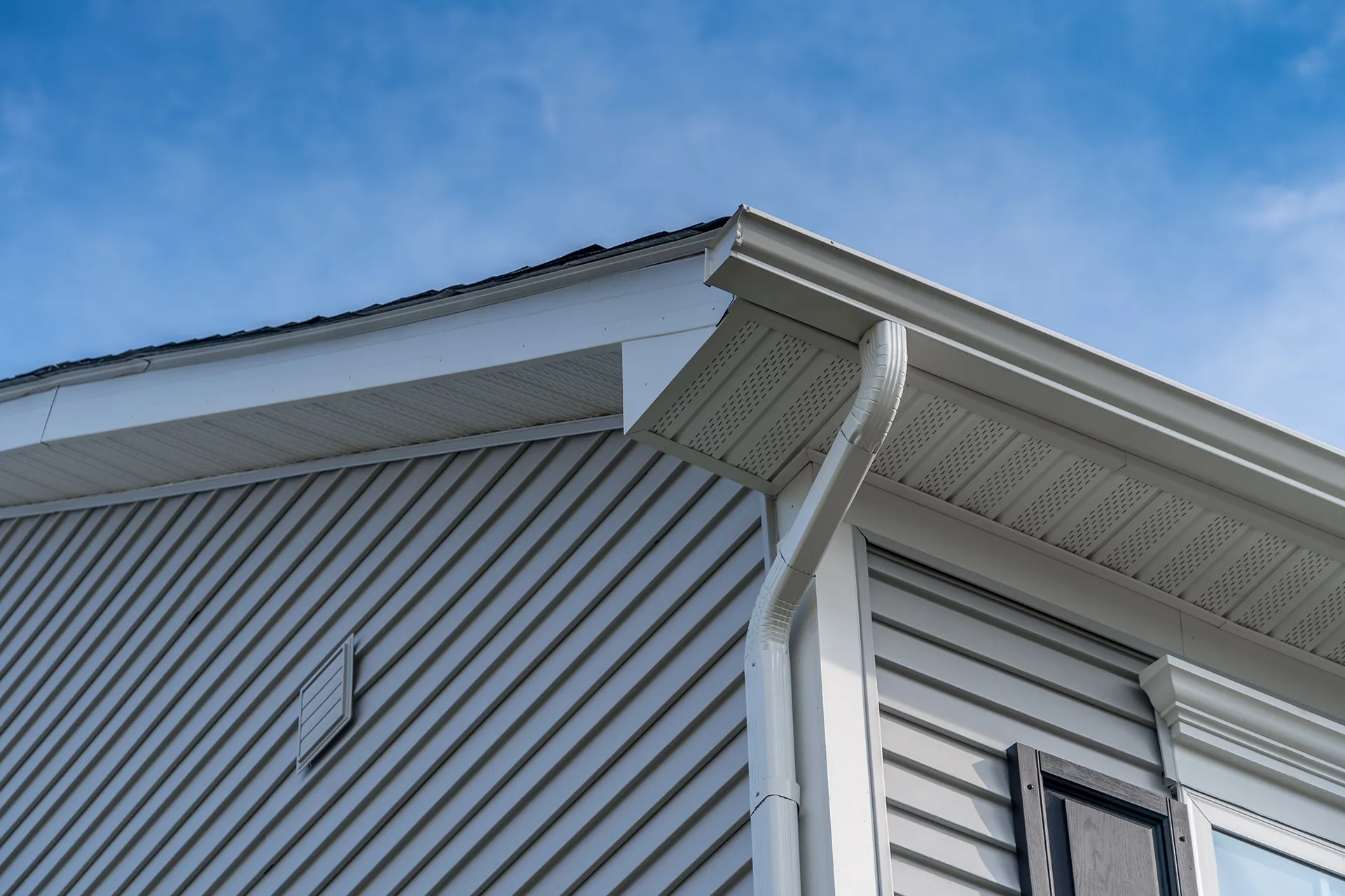 White gutters and downspout on a house with light grey fiber cement siding under a blue sky