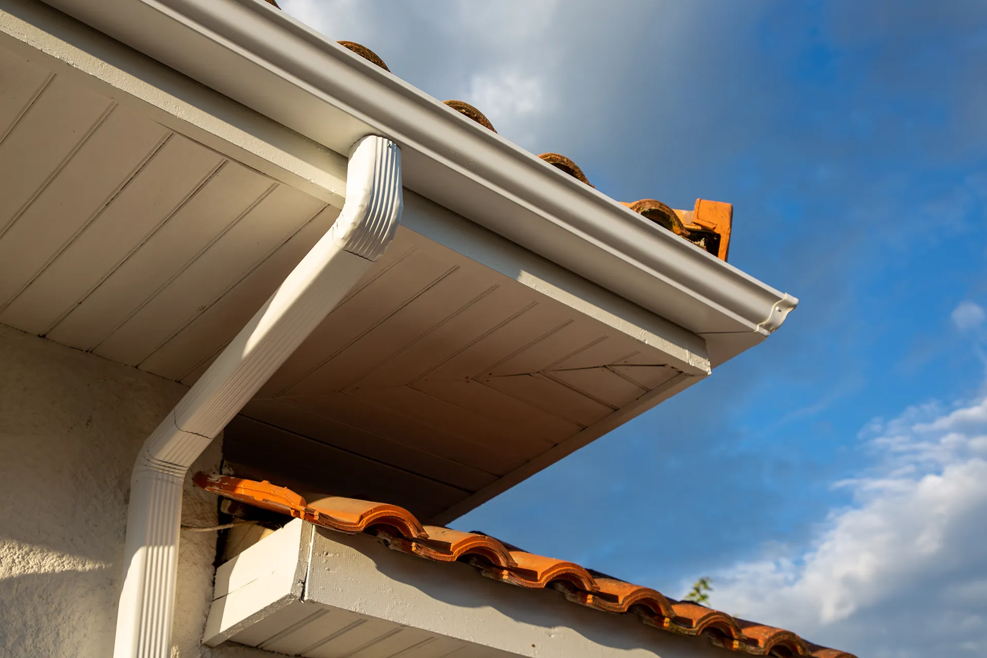 Close-up view of white seamless gutters and downspout on a house with a terracotta tile roof and light-colored soffit, under a partly cloudy blue sky.