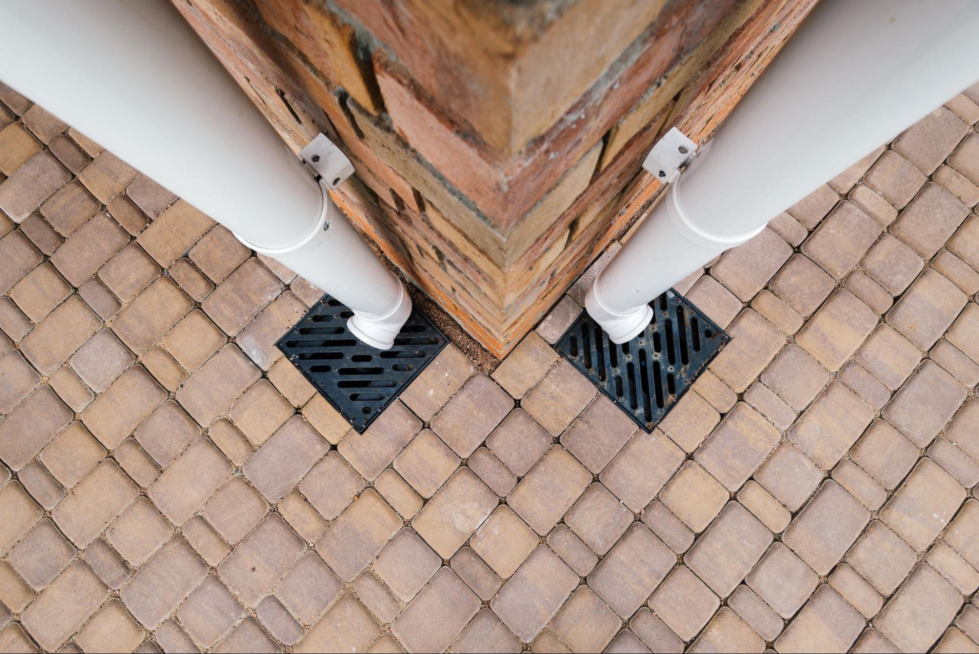 Overhead view of the base of a red brick wall showing two metal downspouts and a small drainage pit