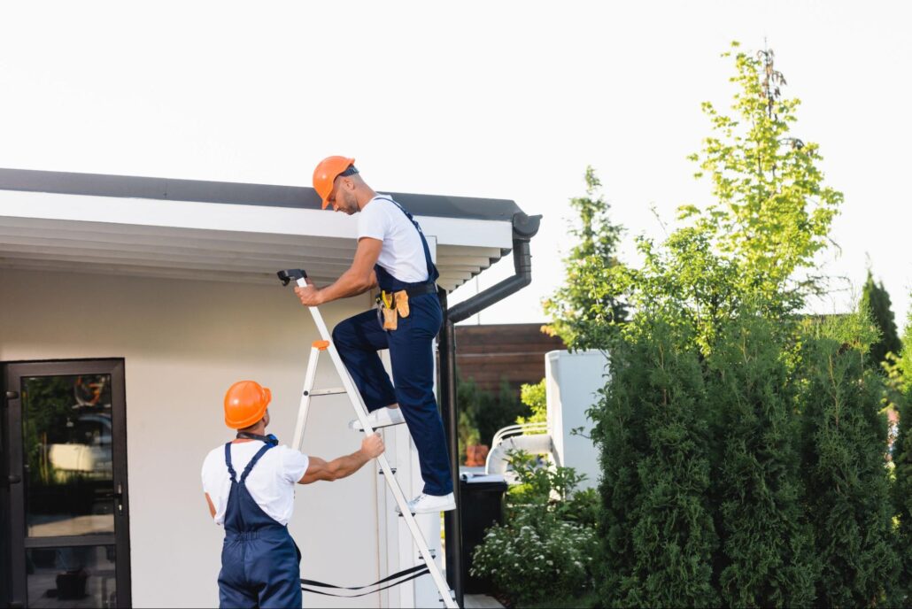 Two workers fixing rain gutters on a home, one on a ladder and one stabilizing it below