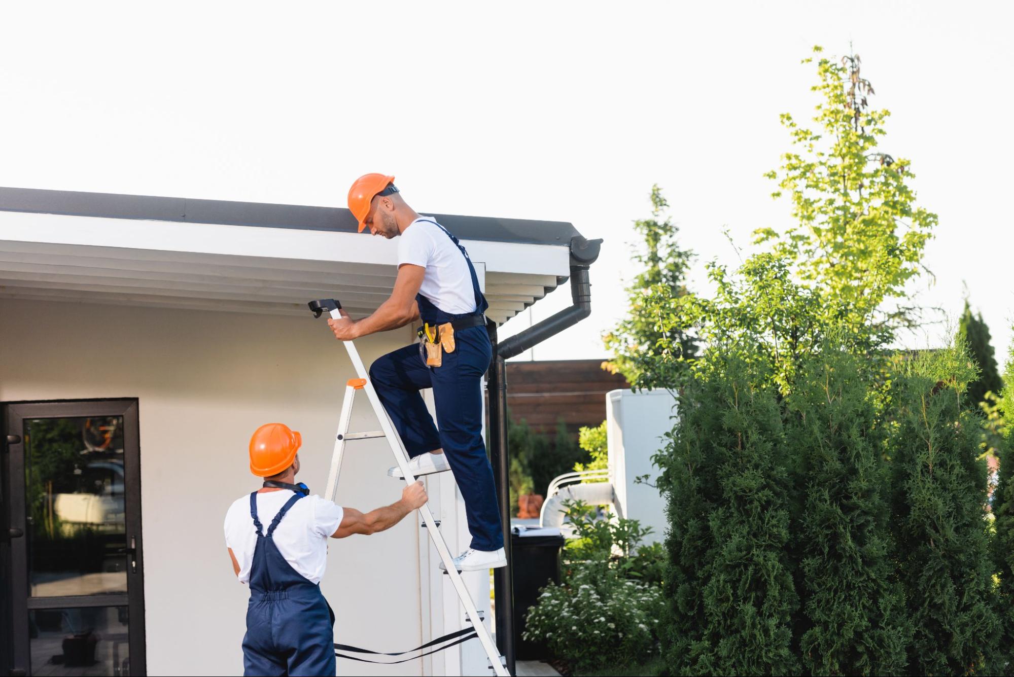 Two workers fixing rain gutters on a home, one on a ladder and one stabilizing it below