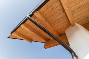 Low-angle view of a gutter downspout attached to wooden roof beams against a clear blue sky