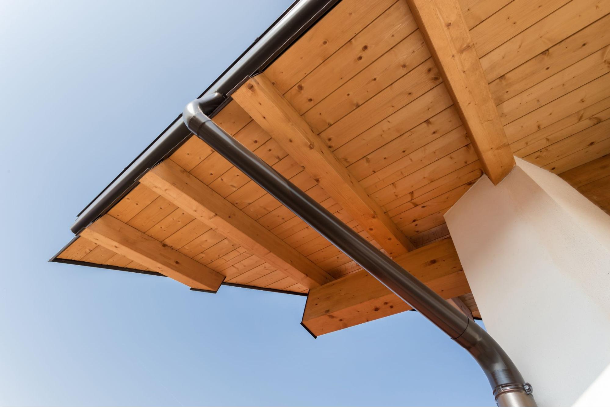 Low-angle view of a gutter downspout attached to wooden roof beams against a clear blue sky